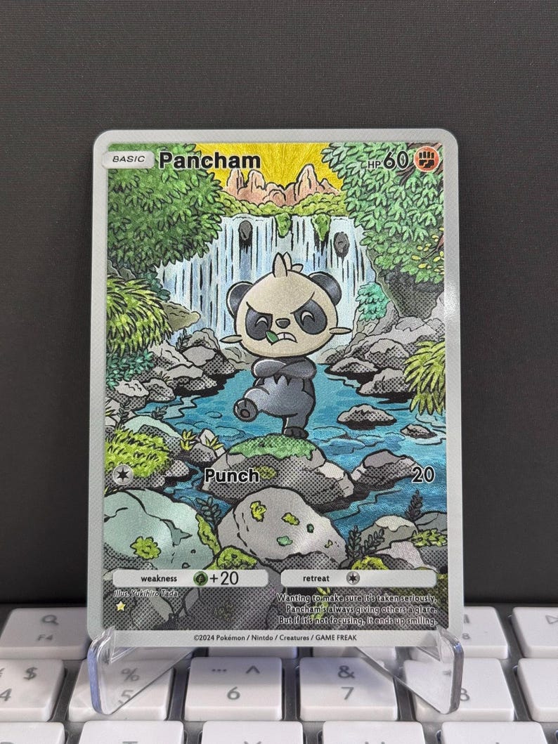 Puede incluir: Una carta coleccionable Pok&eacute;mon de Pancham, que representa una criatura parecida a un panda en un entorno forestal con una cascada. La carta tiene un ataque "Punch" y una debilidad de +20. La carta se muestra en un soporte transparente.