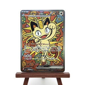 K&ouml;nnte beinhalten: Eine Pok&eacute;mon-Sammelkarte mit einem gelben Meowth-Charakter und japanischem Text, pr&auml;sentiert auf einer kleinen Holzstaffelei. Der Hintergrund der Karte zeigt ein farbenfrohes Design im Glasmalerei-Stil. Sammelobjekt.