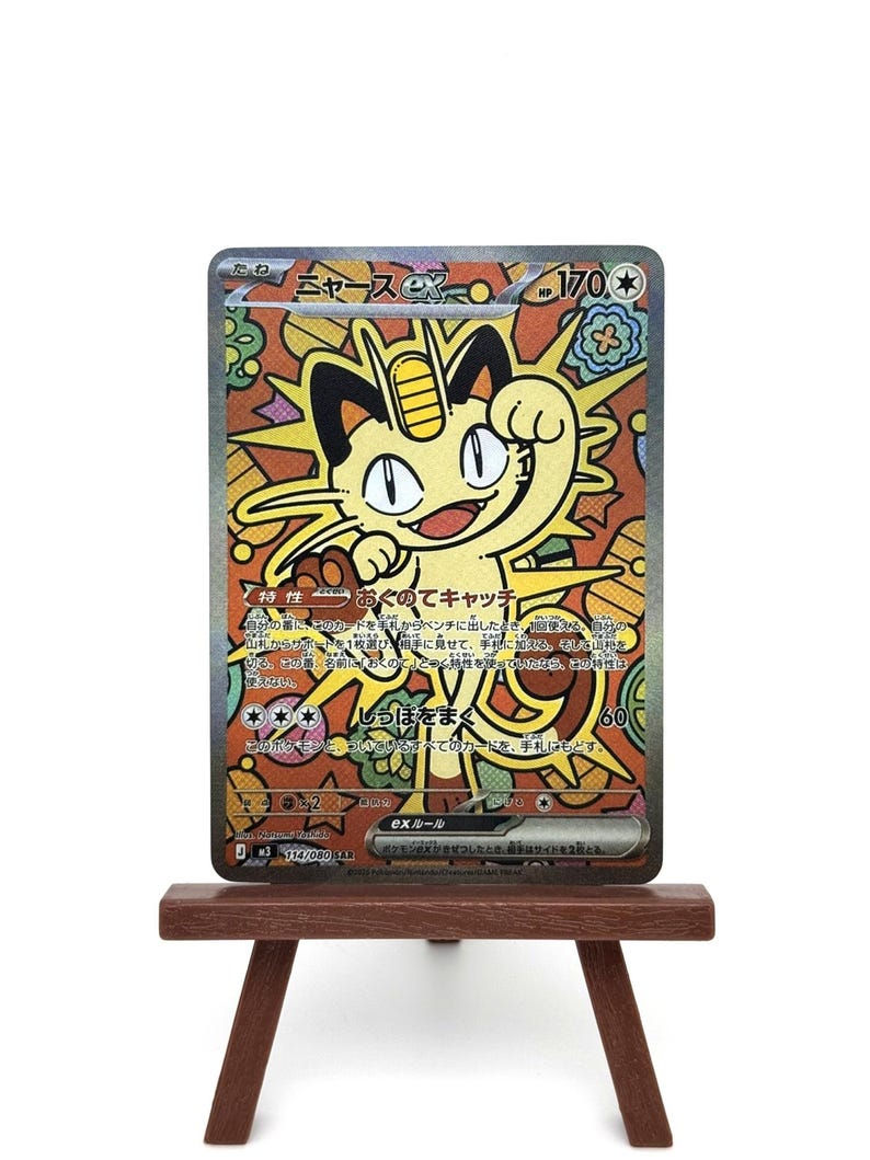 K&ouml;nnte beinhalten: Eine Pok&eacute;mon-Sammelkarte mit einem gelben Meowth-Charakter und japanischem Text, pr&auml;sentiert auf einer kleinen Holzstaffelei. Der Hintergrund der Karte zeigt ein farbenfrohes Design im Glasmalerei-Stil. Sammelobjekt.