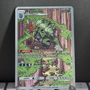 Könnte beinhalten: Holografische Tyranitar Pokémon-Sammelkarte. Die Karte zeigt eine grüne und graue Tyranitar-Illustration vor einem Waldhintergrund. Der Text enthält "STAGE 2 Tyranitar", "HP 160" und Angriffsdetails. Die Karte wird in einem transparenten Kunststoffständer gehalten.