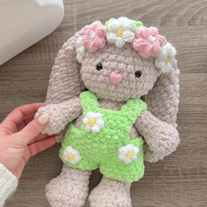 May include: A crocheted plush bunny toy wearing a green and white daisy-patterned overall and a pink and white flower crown.