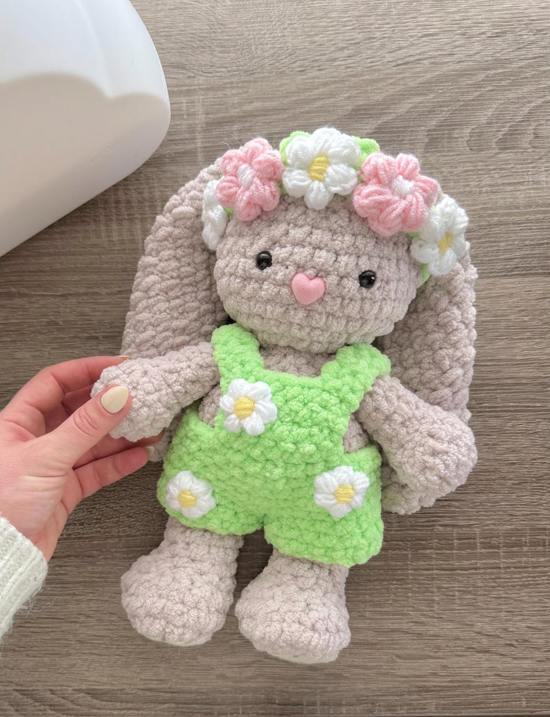 May include: A crocheted plush bunny toy wearing a green and white daisy-patterned overall and a pink and white flower crown.