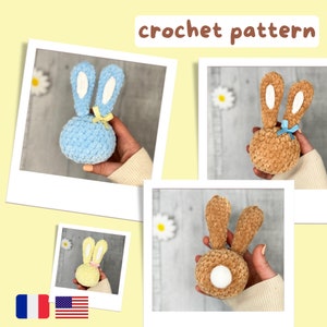 May include: Three crocheted bunny rabbit toys in different colors: blue, brown, and yellow. Each bunny has long ears and a white pom-pom tail. The bunnies are sitting on a light wood surface.