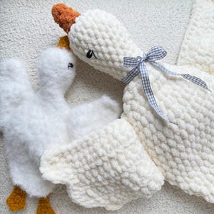 May include: Two white, plush goose-shaped stuffed animals. The larger goose has a brown beak and a small, gray and white checkered bow. The smaller goose has yellow feet and a yellow crown. The plush toys are made of a soft, textured material.