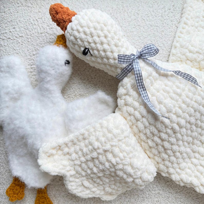 May include: Two white, plush goose-shaped stuffed animals. The larger goose has a brown beak and a small, gray and white checkered bow. The smaller goose has yellow feet and a yellow crown. The plush toys are made of a soft, textured material.