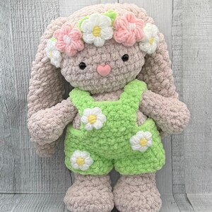 May include: A crocheted bunny toy wearing a flower crown and green overalls with white daisy accents. The bunny has a pink nose and black eyes.
