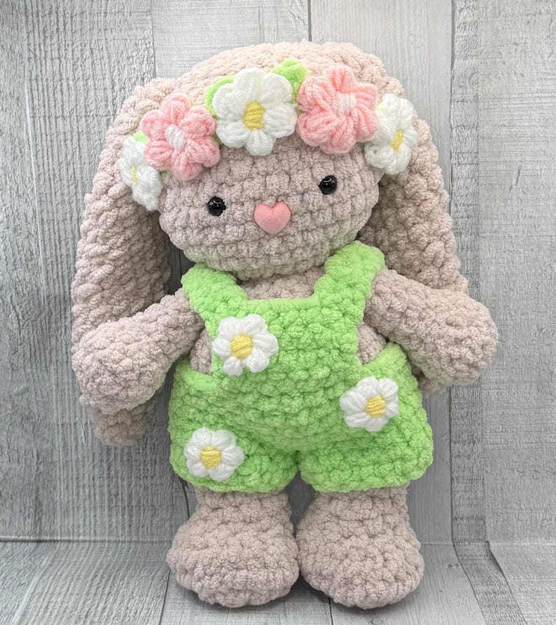 May include: A crocheted bunny toy wearing a flower crown and green overalls with white daisy accents. The bunny has a pink nose and black eyes.