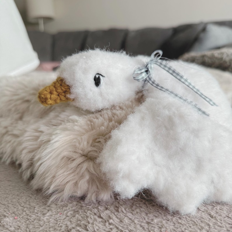 May include: A handmade white plush goose with a yellow beak and black eyes. The goose has a small blue and white checkered ribbon tied around its neck. The goose is lying on a fluffy, light brown surface.