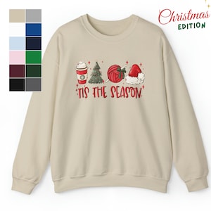 Puede incluir: Una sudadera beige con un diseño navideño. El diseño presenta una taza de café, un árbol de Navidad, una bola de lana y un gorro de Papá Noel. El texto "'Tis the season" está impreso debajo del diseño. El texto "Christmas Edition" está impreso en la esquina superior derecha de la imagen.