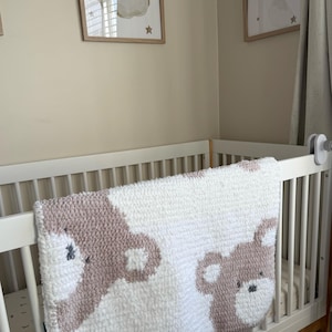 May include: A white and mauve baby blanket with a bear design draped over a white crib. Two framed pictures of bears on clouds hang on the wall above the crib. The blanket is made of a soft, textured material.
