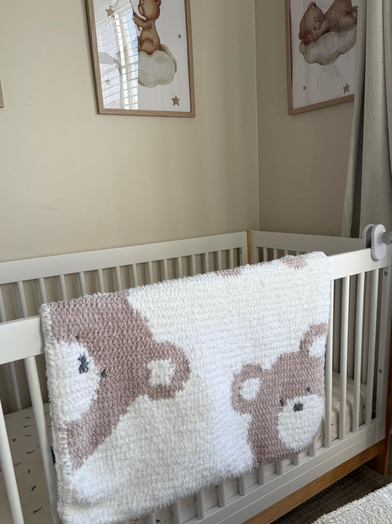 May include: A white and mauve baby blanket with a bear design draped over a white crib. Two framed pictures of bears on clouds hang on the wall above the crib. The blanket is made of a soft, textured material.