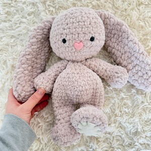 May include: A soft, plush crocheted bunny toy with long ears and a pink heart nose. The bunny is light pink and has black eyes.