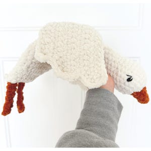 May include: A plush, cream-colored goose toy with outstretched wings and orange feet and beak. The goose has a black eye and is made of a soft, textured material. The toy is held up by a person.