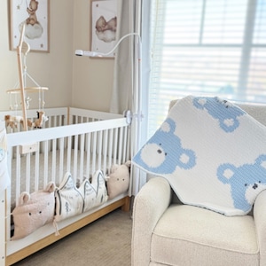 May include: A nursery scene featuring a white crib with decorative pillows and a mobile. A cream-colored armchair has a white and blue bear-patterned blanket draped over it. Two framed pictures hang on the wall.