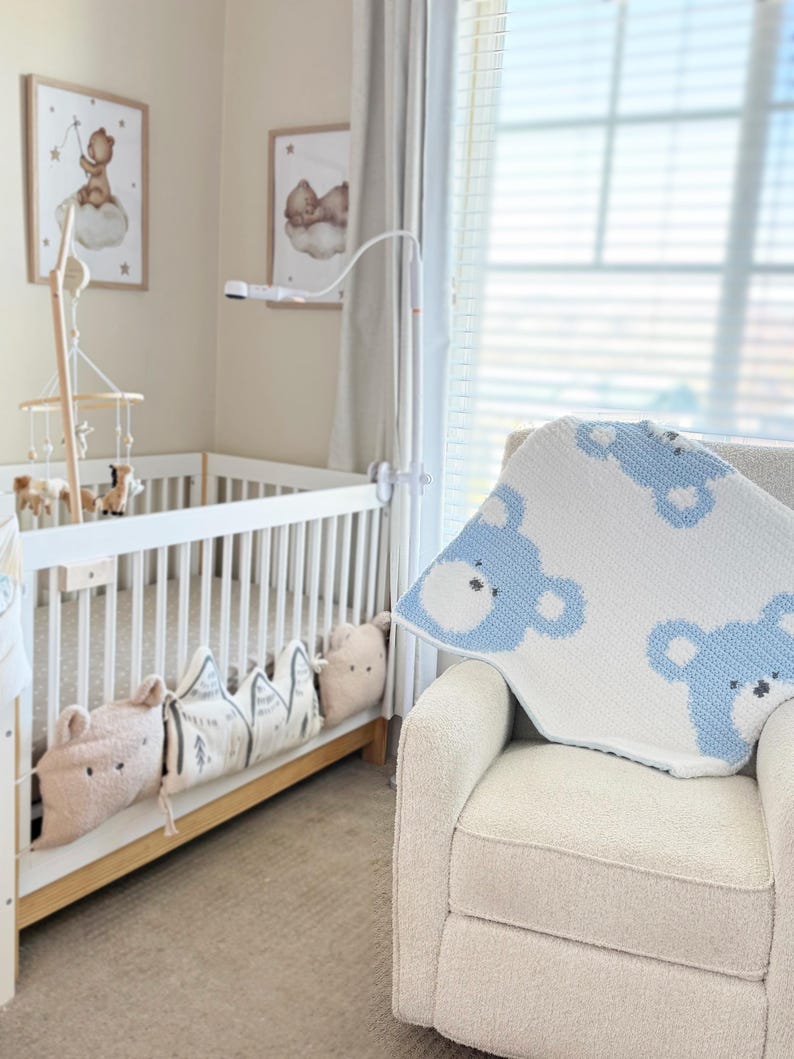 May include: A nursery scene featuring a white crib with decorative pillows and a mobile. A cream-colored armchair has a white and blue bear-patterned blanket draped over it. Two framed pictures hang on the wall.