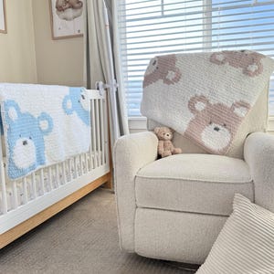 May include: A nursery scene featuring a white crib with a blue bear-patterned blanket and a cream-colored armchair with a pink bear-patterned blanket. A small teddy bear sits on the chair. A framed picture of a bear on a cloud hangs on the wall.