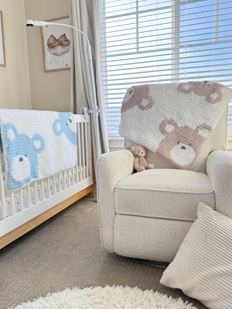 May include: A nursery scene featuring a white crib with a blue bear-patterned blanket and a cream-colored armchair with a pink bear-patterned blanket. A small teddy bear sits on the chair. A framed picture of a bear on a cloud hangs on the wall.