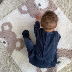 May include: A handmade baby blanket featuring a repeating bear design in white and taupe. The blanket is made of a soft, textured material. A baby in a navy blue onesie is lying on the blanket.