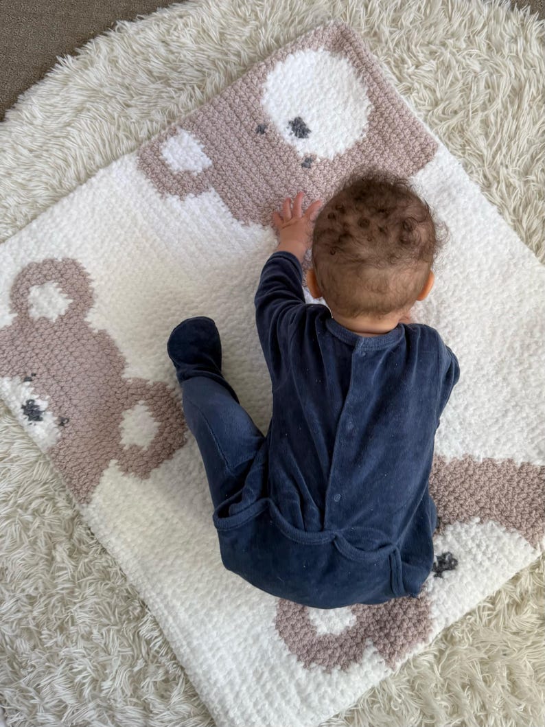 May include: A handmade baby blanket featuring a repeating bear design in white and taupe. The blanket is made of a soft, textured material. A baby in a navy blue onesie is lying on the blanket.