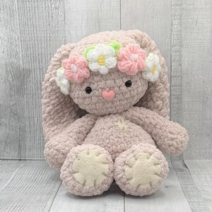 May include: A pink crocheted bunny with long floppy ears wearing a flower crown. The crown is made of pink, white, and green crocheted flowers.
