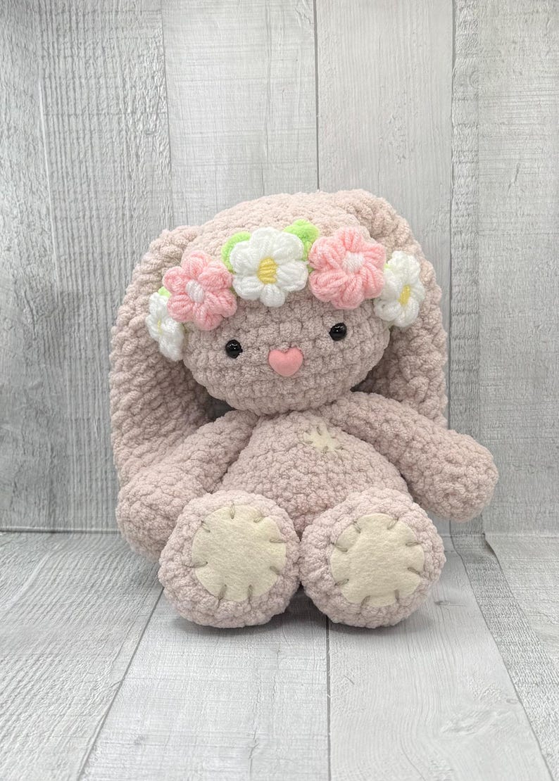 May include: A pink crocheted bunny with long floppy ears wearing a flower crown. The crown is made of pink, white, and green crocheted flowers.