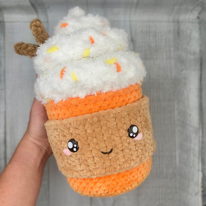 May include: A crocheted plush toy shaped like a pumpkin spice latte. The toy has a white fluffy top with orange and yellow sprinkles, an orange crocheted cup, and a brown crocheted rim. The toy has a cute cartoon face with black eyes and a smile.