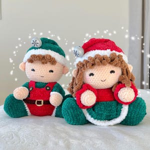 K&ouml;nnte beinhalten: Zwei geh&auml;kelte Elfenpuppen, eine tr&auml;gt ein gr&uuml;nes und rotes Outfit und die andere ein gr&uuml;nes und rotes Kleid mit wei&szlig;em Besatz. Beide Puppen haben braunes Haar und l&auml;chelnde Gesichter.
