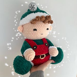 K&ouml;nnte beinhalten: Eine geh&auml;kelte Elfenpuppe mit einem gr&uuml;n-roten Outfit, braunen Haaren und einer wei&szlig;en M&uuml;tze mit einem silbernen Schneeflocken-Anh&auml;nger. Der Elf l&auml;chelt freundlich.