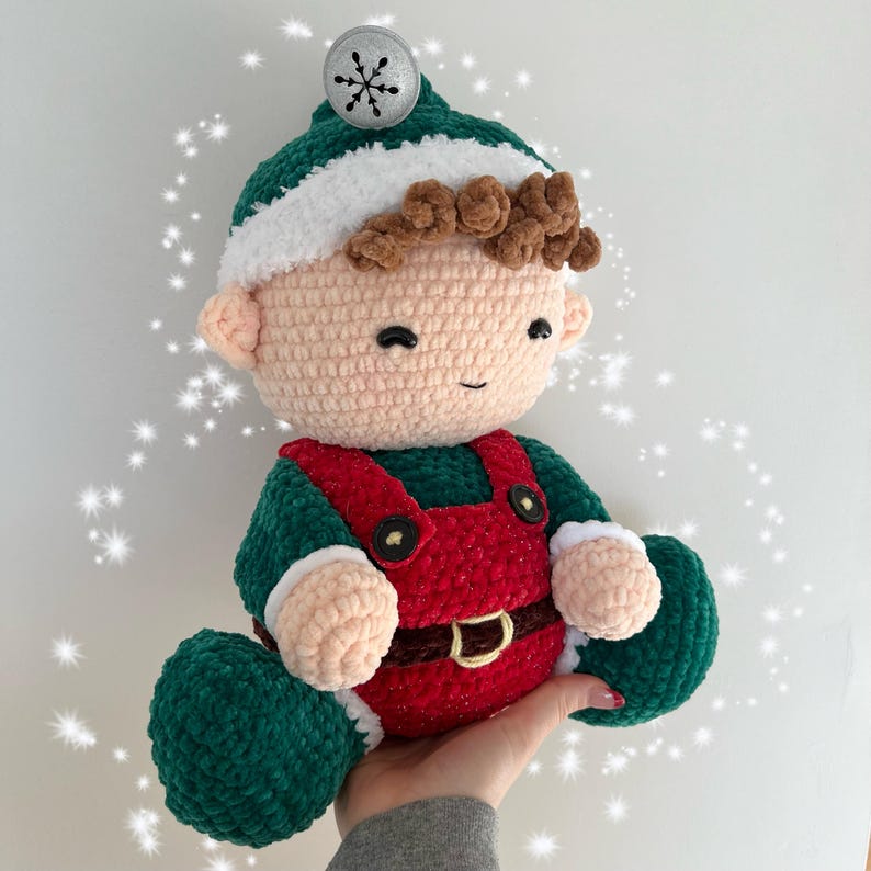 K&ouml;nnte beinhalten: Eine geh&auml;kelte Elfenpuppe mit einem gr&uuml;n-roten Outfit, braunen Haaren und einer wei&szlig;en M&uuml;tze mit einem silbernen Schneeflocken-Anh&auml;nger. Der Elf l&auml;chelt freundlich.