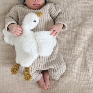 May include: A baby wearing a beige knit outfit and a blue cap, holding a white plush goose toy with yellow feet and beak. The goose has black eyes. The baby is lying on a beige textured blanket.