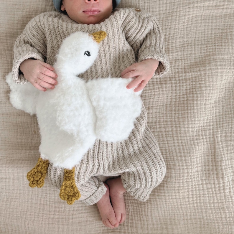 May include: A baby wearing a beige knit outfit and a blue cap, holding a white plush goose toy with yellow feet and beak. The goose has black eyes. The baby is lying on a beige textured blanket.