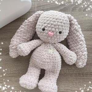 May include: A soft, plush, light purple crocheted bunny toy with a pink nose and black eyes. The bunny is lying on a wooden surface.