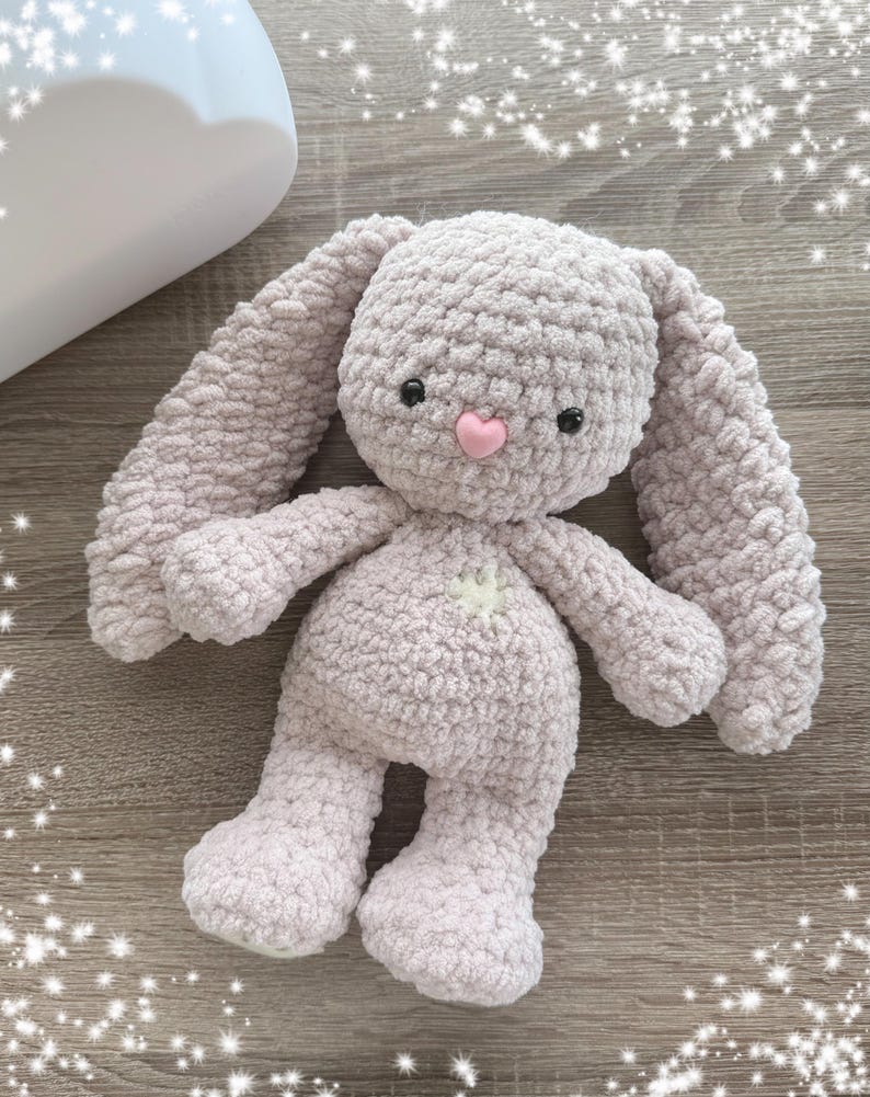 May include: A soft, plush, light purple crocheted bunny toy with a pink nose and black eyes. The bunny is lying on a wooden surface.