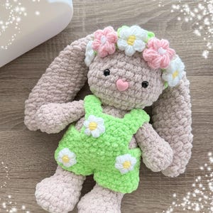 May include: A crocheted bunny toy wearing a green and white floral overall and a pink and white flower crown.