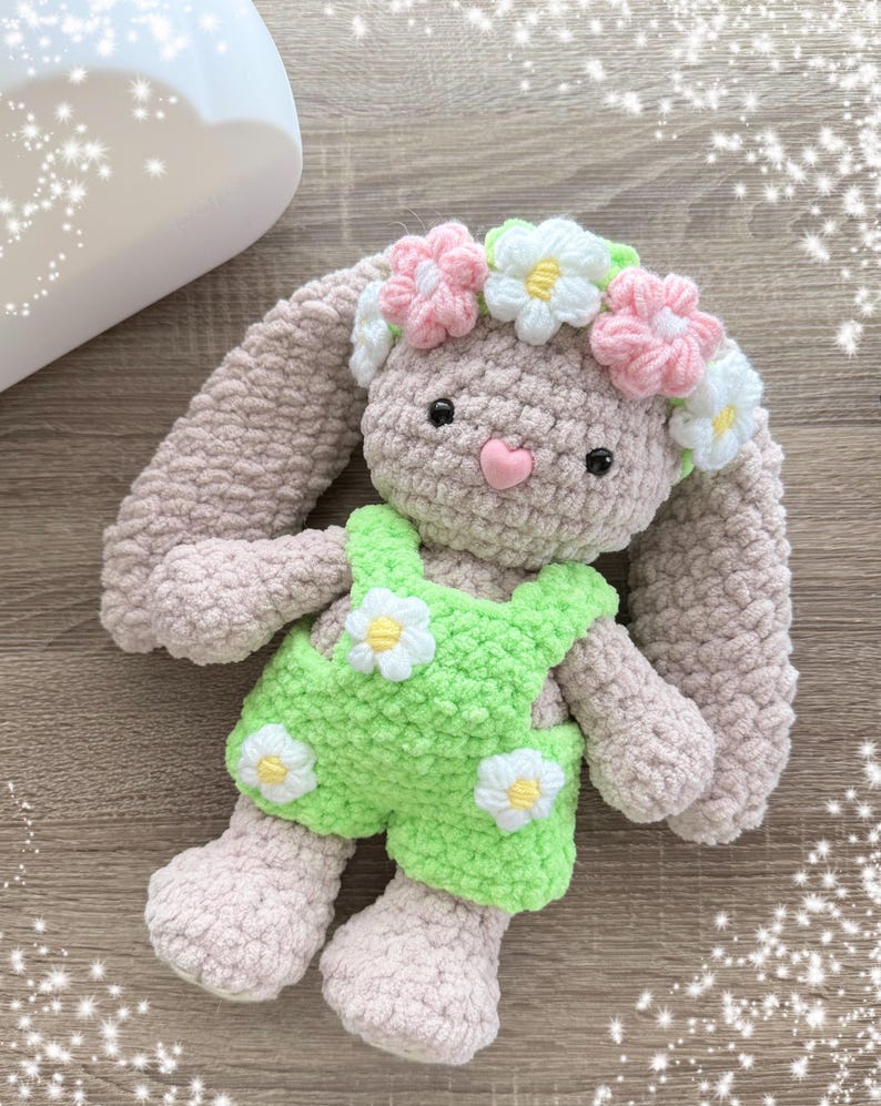 May include: A crocheted bunny toy wearing a green and white floral overall and a pink and white flower crown.