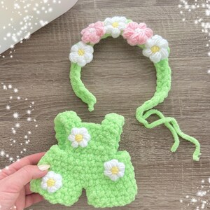 May include: A green crocheted outfit with white daisy embellishments and a matching headband with pink and white crocheted flowers.