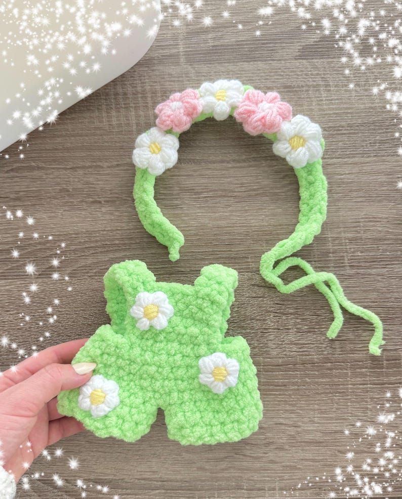 May include: A green crocheted outfit with white daisy embellishments and a matching headband with pink and white crocheted flowers.
