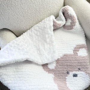 May include: A white and pink crochet baby blanket with a teddy bear face design. The blanket is folded over a white armchair. The teddy bear face has gray accents for the eyes and nose. The blanket is made of soft, fluffy yarn.