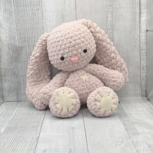 May include: A soft, pink crocheted bunny rabbit toy with black eyes and a pink nose. The bunny has long floppy ears and is sitting on a light wood background.