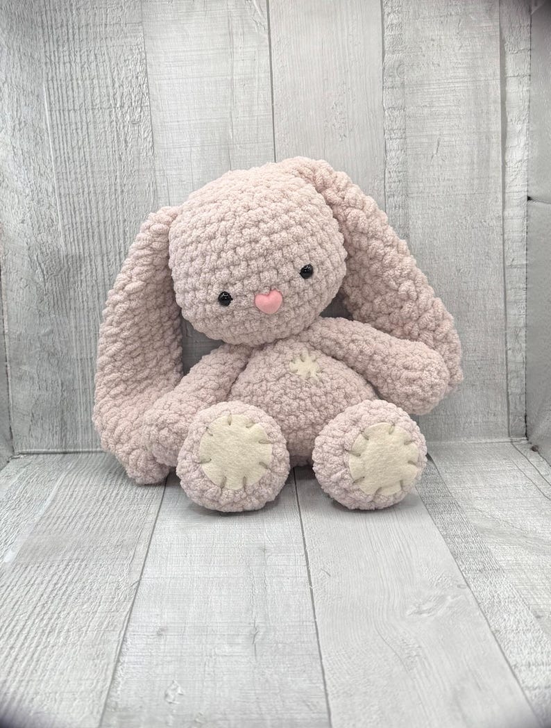 May include: A soft, pink crocheted bunny rabbit toy with black eyes and a pink nose. The bunny has long floppy ears and is sitting on a light wood background.