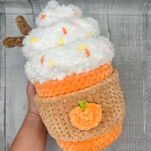 May include: A crocheted pumpkin spice latte plush toy with a white whipped cream top, orange and brown cup, and a small orange pumpkin on the side.