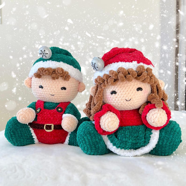 K&ouml;nnte beinhalten: Zwei geh&auml;kelte Weihnachts-Elfenpuppen, eine mit gr&uuml;nem Hut und roter Latzhose, die andere mit rotem Hut und gr&uuml;nem Kleid. Beide Puppen haben braunes Haar und l&auml;chelnde Gesichter.