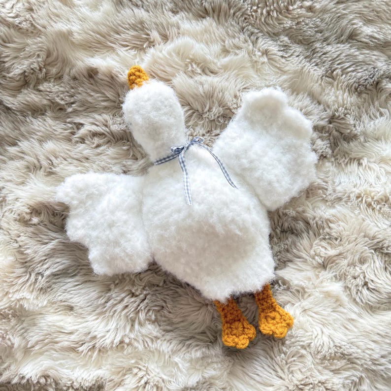 May include: A fluffy white goose-shaped plush toy with outstretched wings. The toy has a yellow beak and feet, and a blue and white checkered ribbon around its neck. The plush is lying on a soft, textured, light-colored surface.