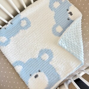 May include: A handmade baby blanket featuring a white and light blue bear design. The blanket is crocheted with a soft, textured yarn and has a light blue, dimpled backing. The bear's face is detailed with gray accents.
