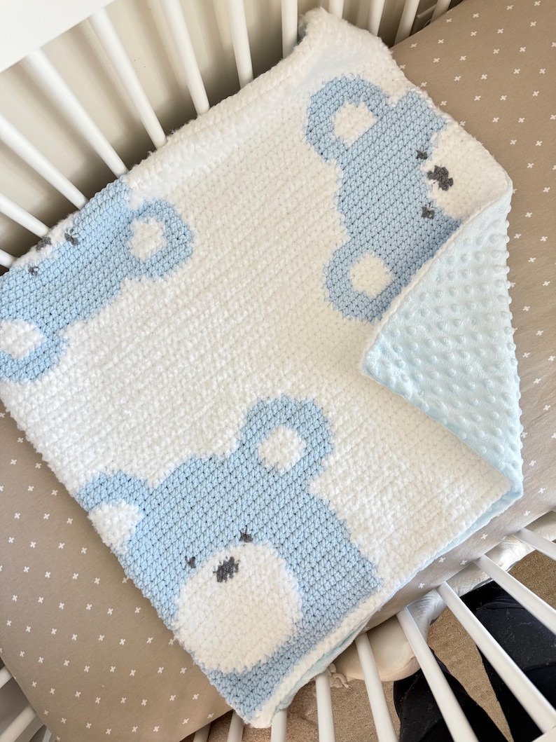 May include: A handmade baby blanket featuring a white and light blue bear design. The blanket is crocheted with a soft, textured yarn and has a light blue, dimpled backing. The bear's face is detailed with gray accents.