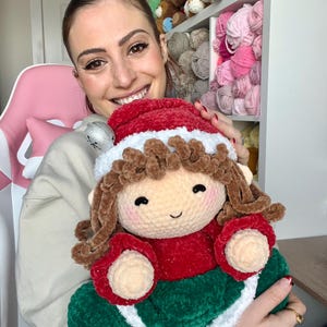 K&ouml;nnte beinhalten: Eine geh&auml;kelte Elfenpuppe, die ein rot-gr&uuml;nes Kleid mit wei&szlig;em Besatz tr&auml;gt. Die Puppe hat braunes Haar und ein l&auml;chelndes Gesicht.