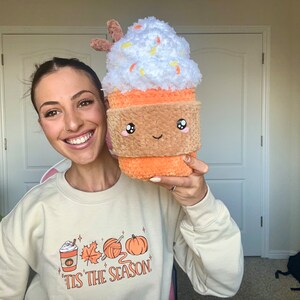 May include: A woman is holding a plush toy shaped like a pumpkin spice latte. The toy has a white whipped cream top with orange sprinkles, a brown rim, and a smiling face. The woman is wearing a cream colored sweatshirt with the text "'Tis the season" and a graphic of a coffee cup, yarn, and a pumpkin.