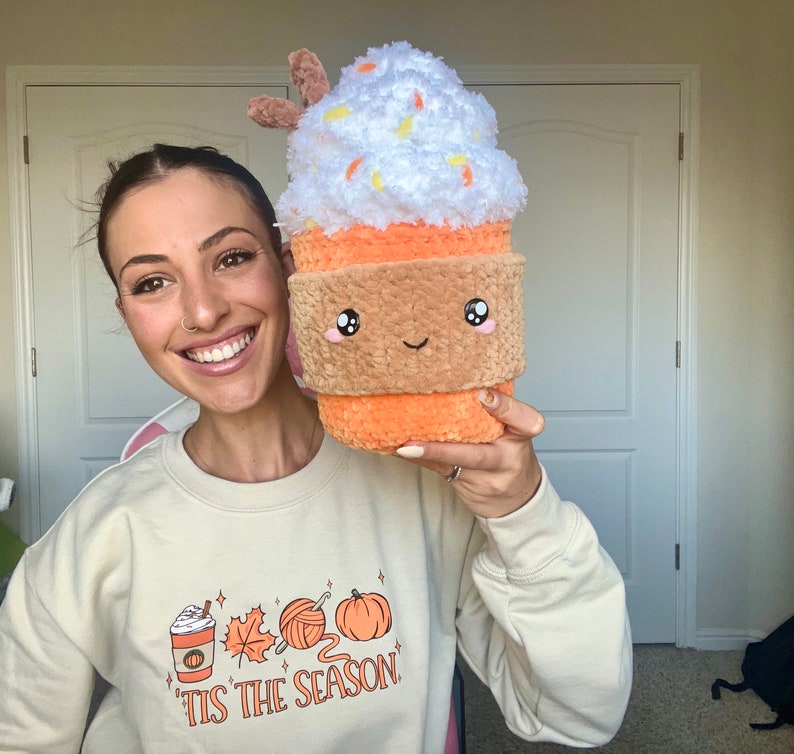 May include: A woman is holding a plush toy shaped like a pumpkin spice latte. The toy has a white whipped cream top with orange sprinkles, a brown rim, and a smiling face. The woman is wearing a cream colored sweatshirt with the text "'Tis the season" and a graphic of a coffee cup, yarn, and a pumpkin.