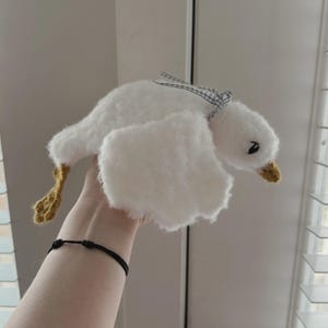 May include: A handmade white plush goose with a blue and white checkered ribbon tied around its neck. The goose has yellow feet and a yellow beak. The wings are outstretched, giving the impression of flight. The plush toy is held up in front of a window.