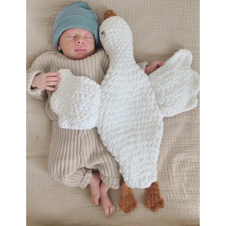 May include: A sleeping baby wearing a light blue beanie and a beige knit outfit, lying next to a large white goose-shaped plush toy with brown feet and a brown beak. The background is a light beige fabric.